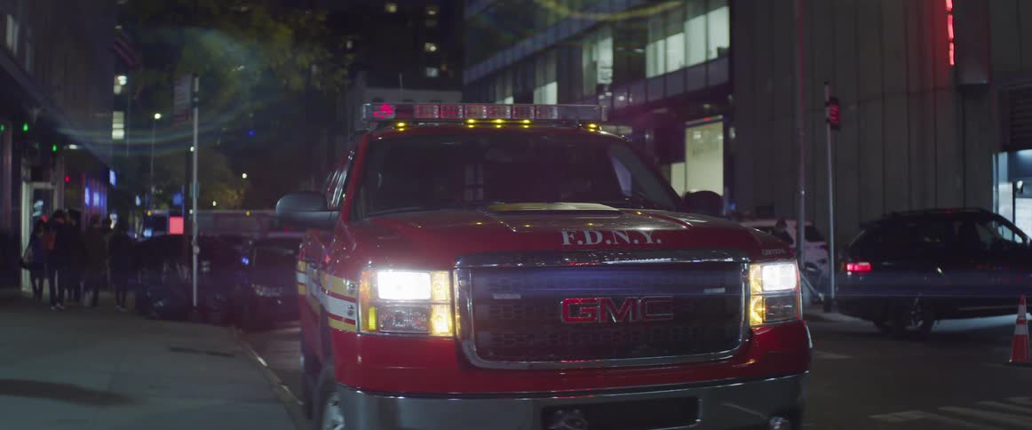 A fire department sport utility vehicle is parked with its lights on next to a city curb at night as pedestrians pass by