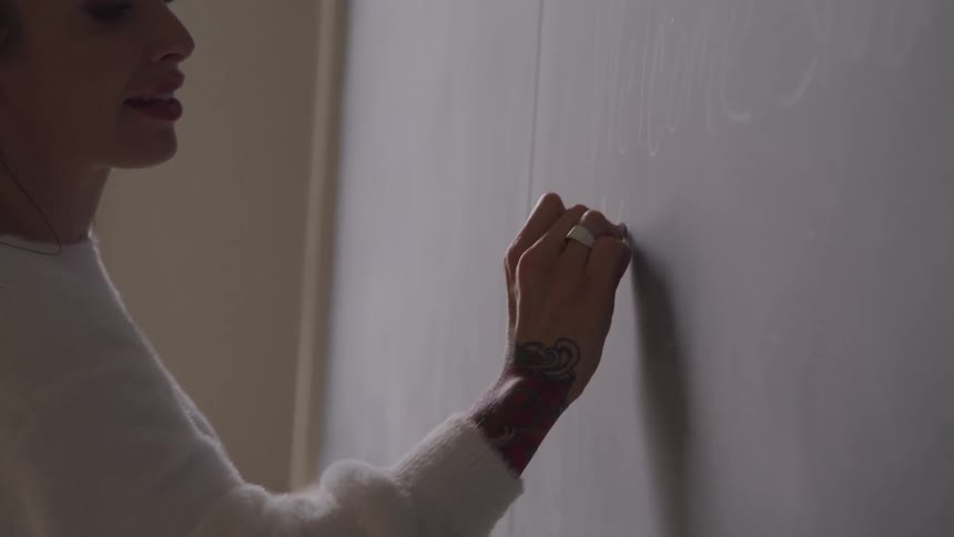 A woman with visible tattoos on her arm writes on a chalkboard with chalk