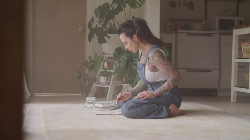 A woman sits and paints on the floor of her apartment in front of a large window