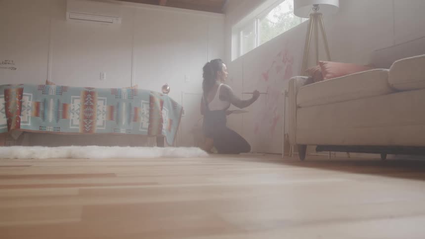 Woman kneeling on the floor of a room paints a wall with a paintbrush