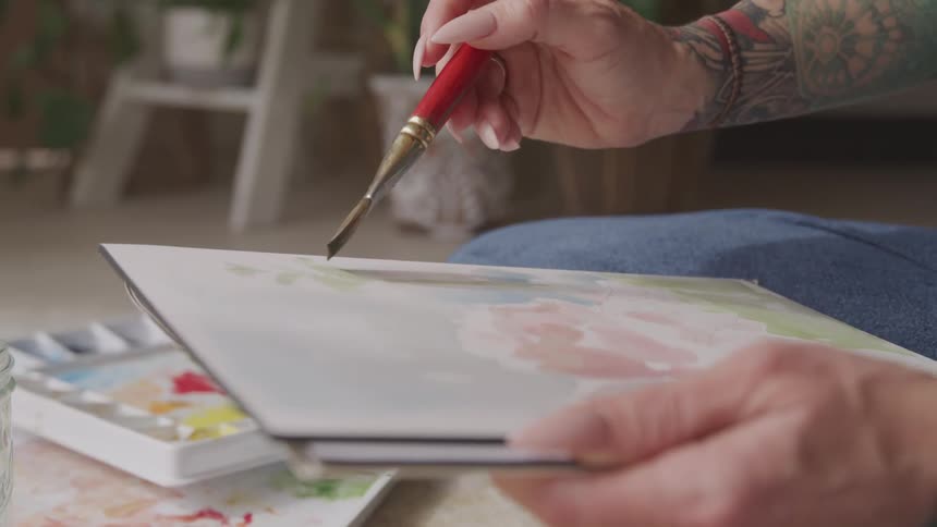 A tattooed woman painting while sitting cross legged on the floor