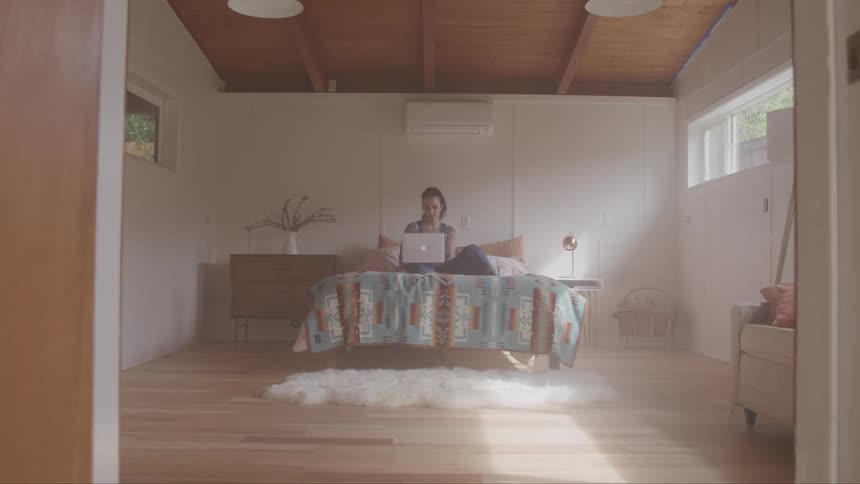 Tattooed woman sitting on the bed uses her laptop in the bedroom