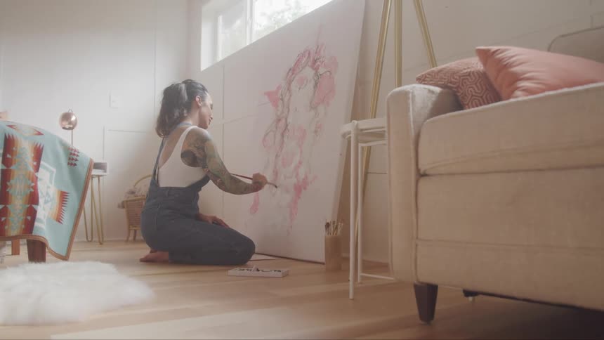 A woman kneels on the floor painting a canvas