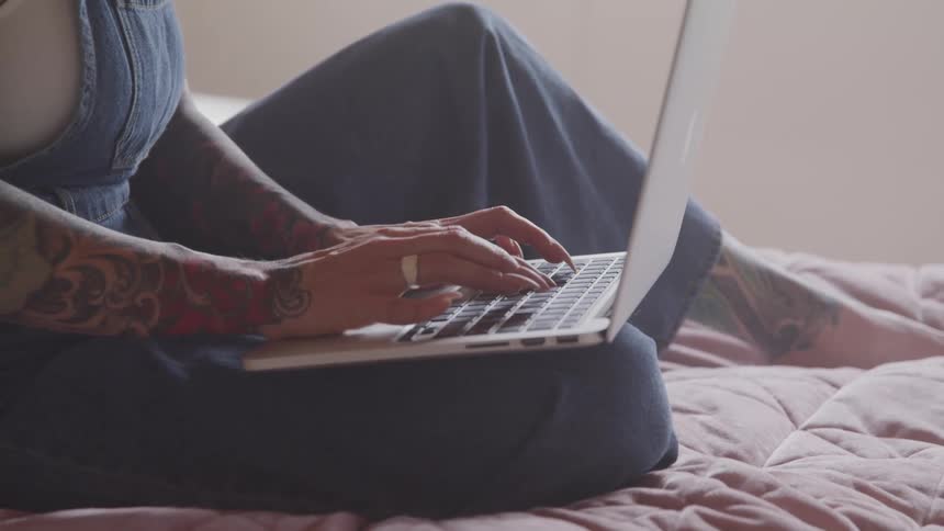 Tattooed woman sitting on the bed uses a laptop in her room