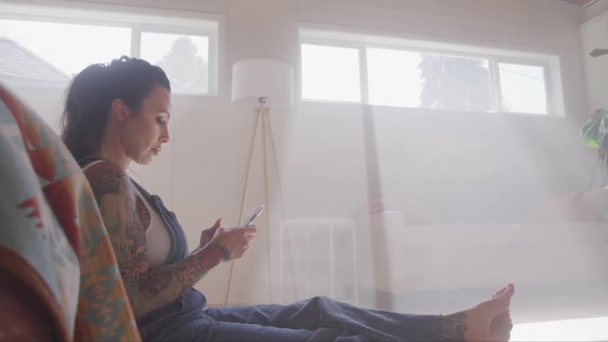 Tattooed woman sitting on the ground uses a smartphone in the living room at home