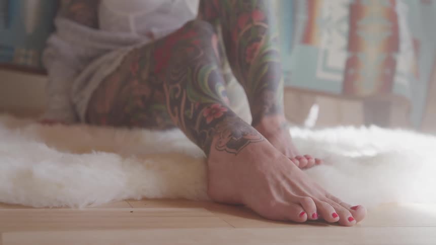 A woman with arms and legs tattooed sits on a white furry rug wriggling her toes