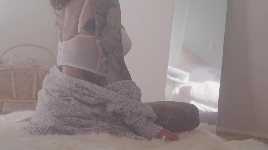 A woman covered in tattoos sits on a fur comforter in her bedroom with only a bra on