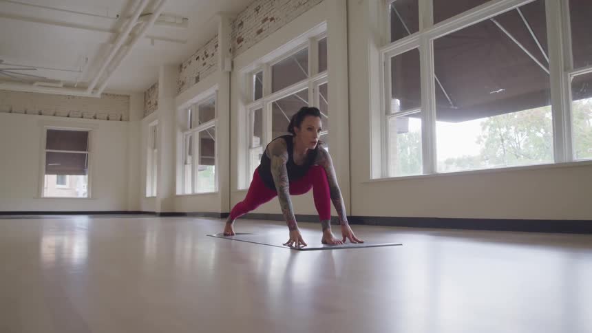 A woman with full sleeve tattoos stretches and holds yoga poses in a large empty studio