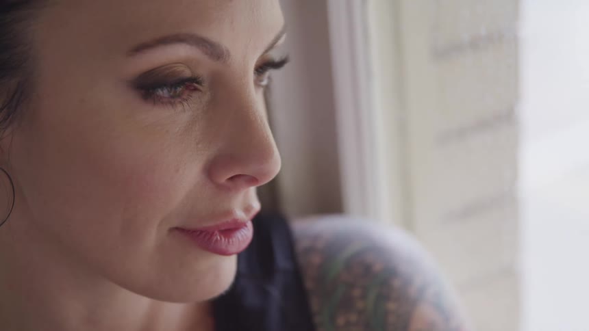 A woman with arm tattoos leans on a window looking out with a small smile on her face