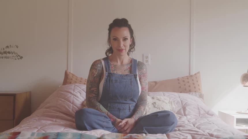 Woman in overalls with sleeve tattoos sitting on bed