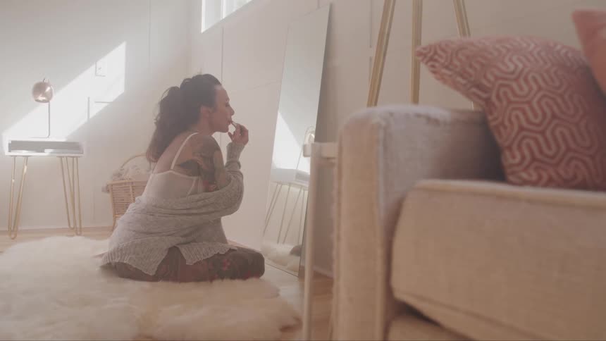 Woman sits on floor rug in living room applying lipstick in front of mirror