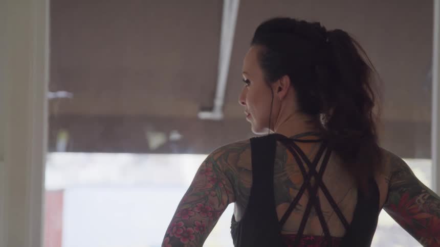 A woman wearing a sports bra and with tattoos covering her back stands under a pier