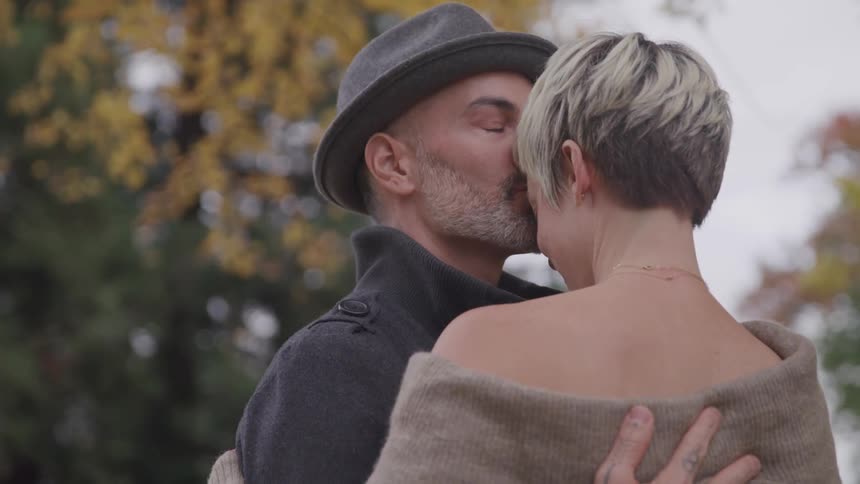 Male and female couple embrace one another beneath the trees in the fall and the female smiles when the male leans in to kiss her on the forehead
