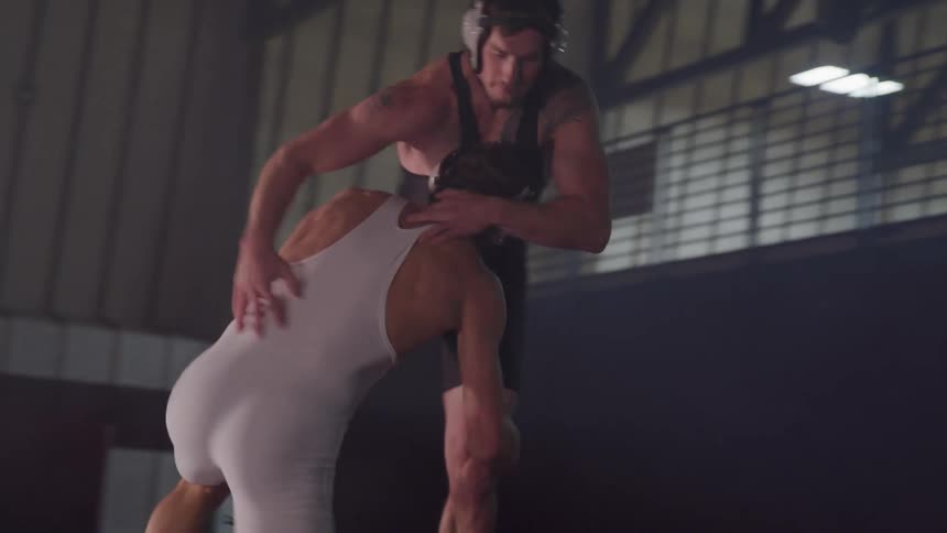Two male high school wrestlers wearing headgear are practicing wrestling moves together in a gymnasium