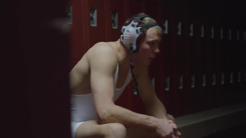 A teenager in wrestling gear sits in a locker room