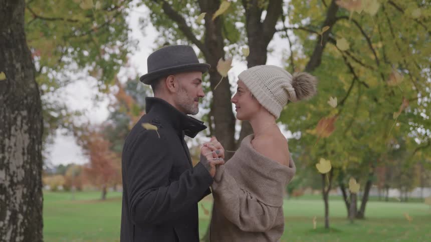A man and woman face each other and hold hands in a public park with leaves showering over them