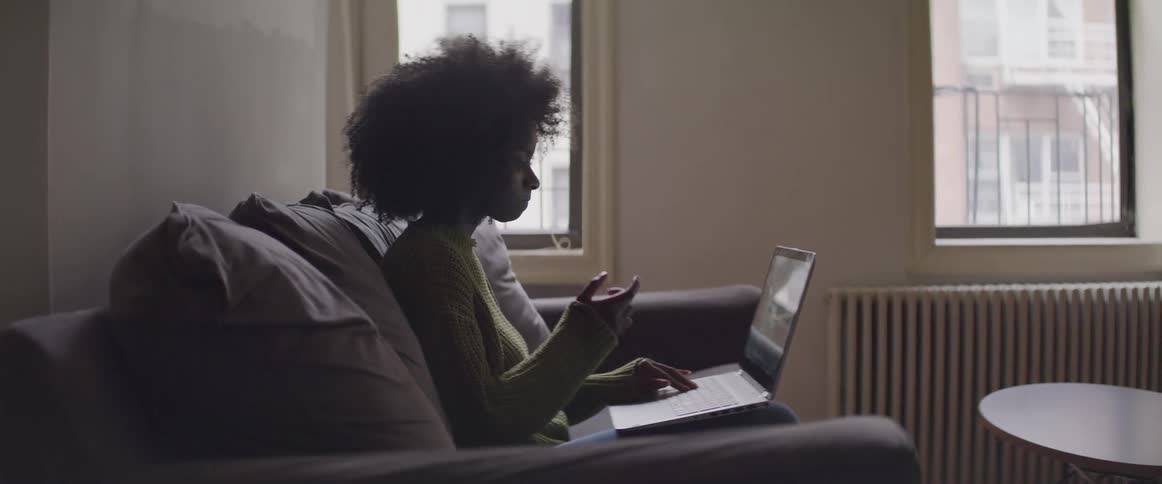 A woman sits on a couch looking at a laptop and places her hand on her face