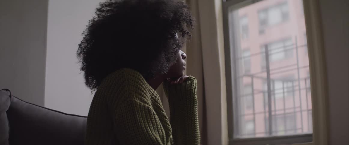 Woman in apartment sits in chair while looking outside window