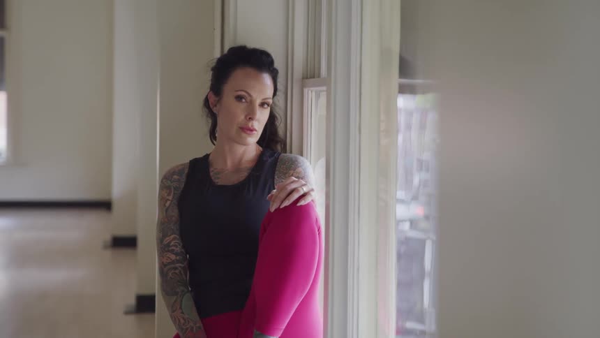 A woman with tattoos sitting by a window and staring