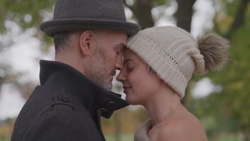 Romantic couple standing close at a park with eyes closed as leaves fall around them