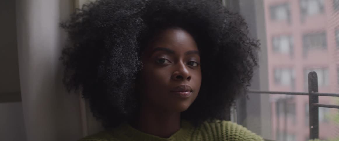 Black woman sits near a window and turns her head to look at the camera