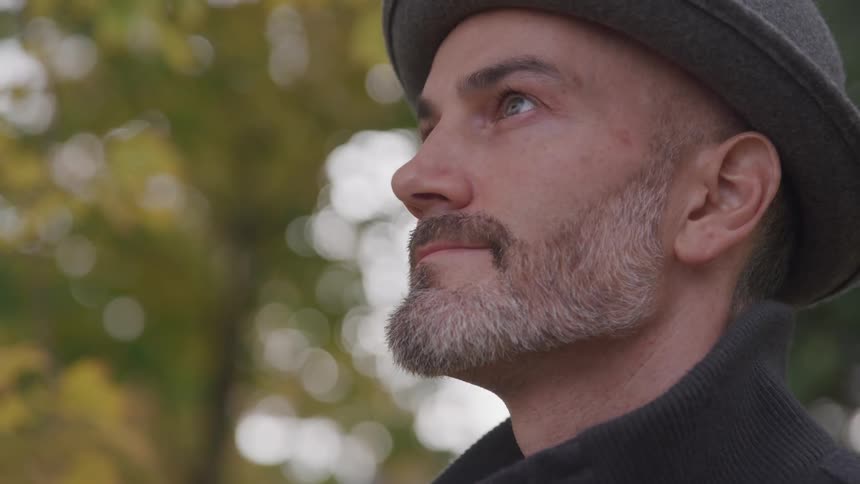 A man with a graying beard wearing a hat looking up at the trees and sky