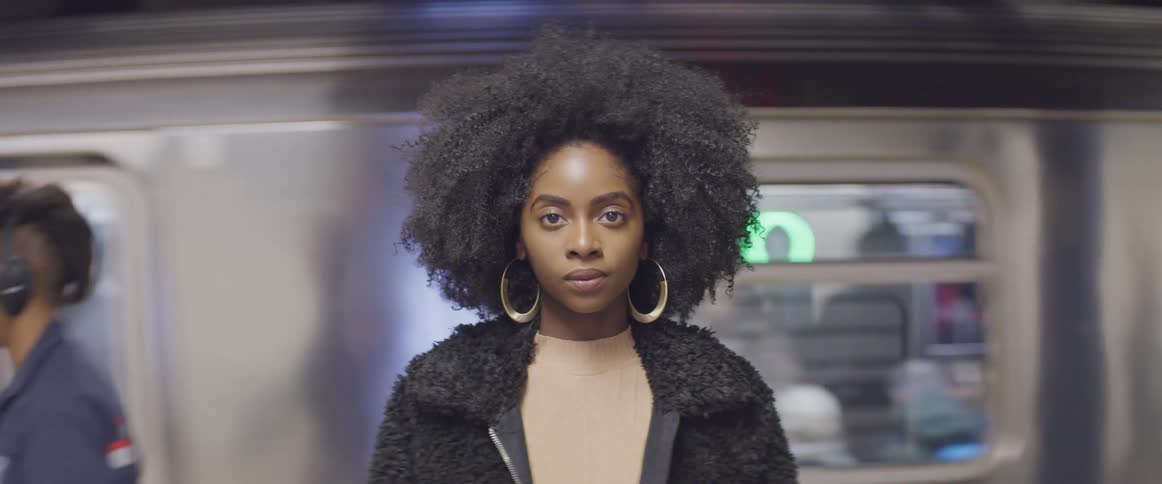 Female with large hoop earring stands still and stares straight ahead as a male and the subway train behind her continue to move