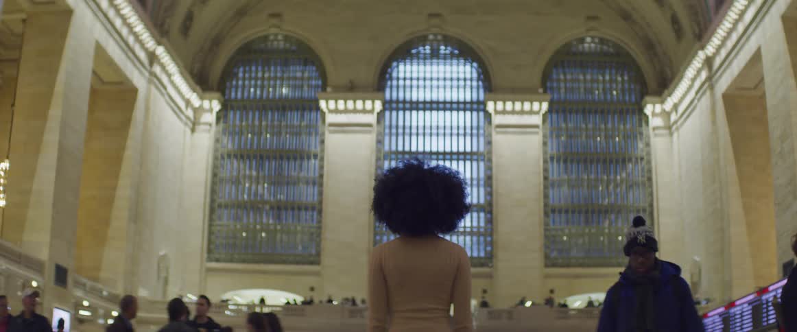 A woman stands and stares at arched windows in a busy train depot as people pass by