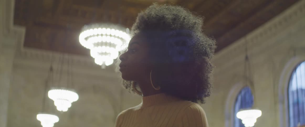 A woman with an afro and gold hoop earrings looks up and around while standing in a room with chandeliers