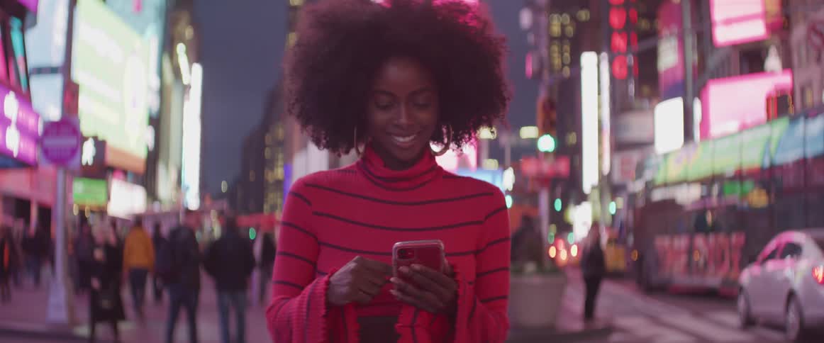 A woman stands next to a busy road downtown and laughs while using her smart phone