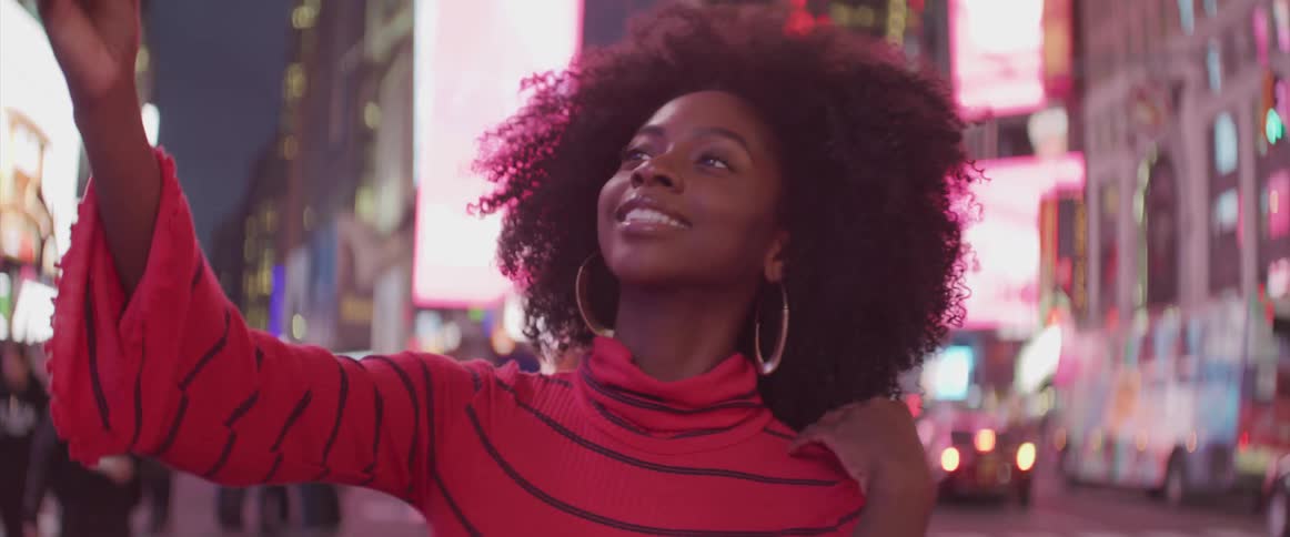 A young black woman posing for selfies with the colorful city lights behind her at night