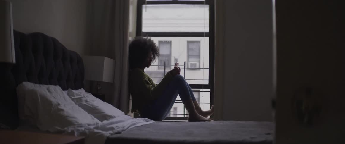 A black woman sits by a window in a bedroom and uses a smartphone