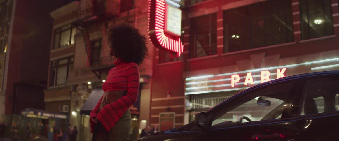 A woman stands beside a car in a large city looking like she is waiting for someone