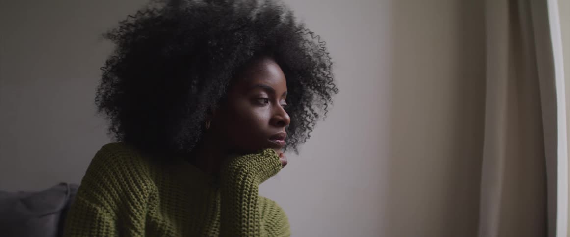 Side view of a curly haired black woman looking out the window in the living room