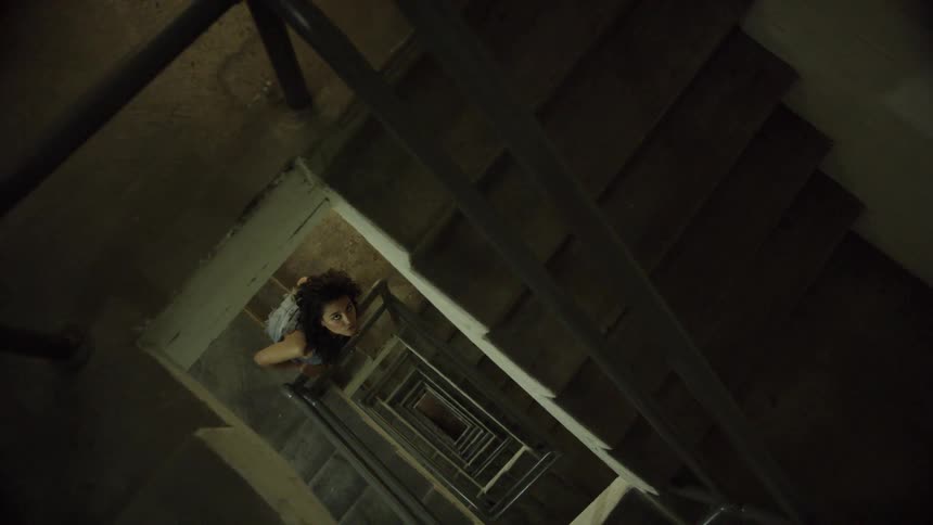 A white woman with dark hair peers up through the opening in a stairwell