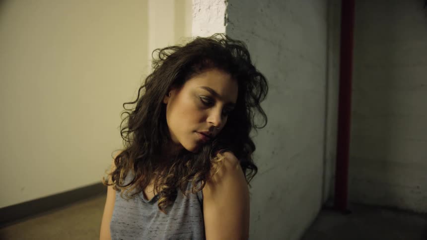A woman with curly black hair poses against a wall next to a hallway