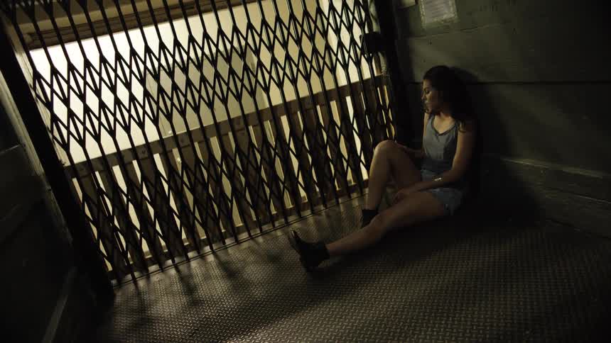 A woman sits on the floor of a large old fashioned elevator and watches the floors as she passes them through the gates