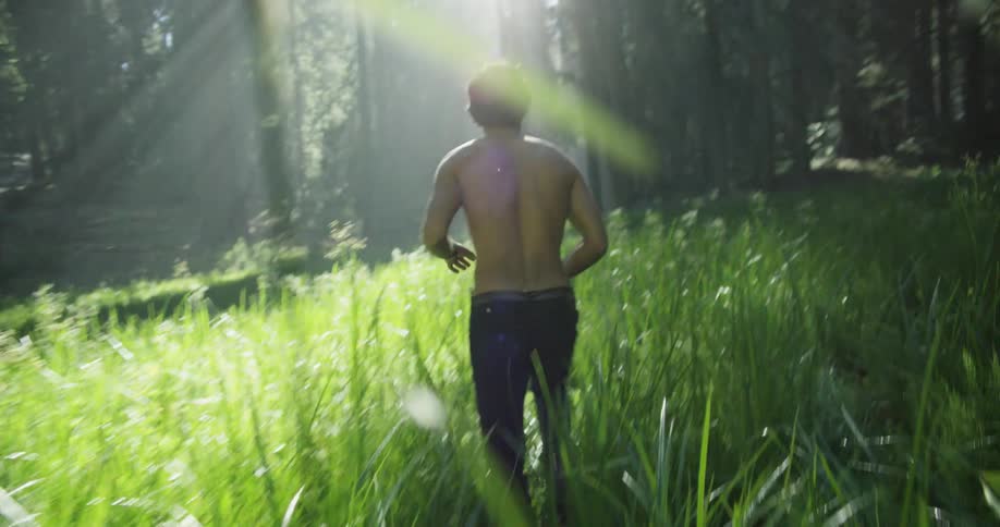 Shirtless male runs through the tall grass towards the woods as the sun beams through the trees