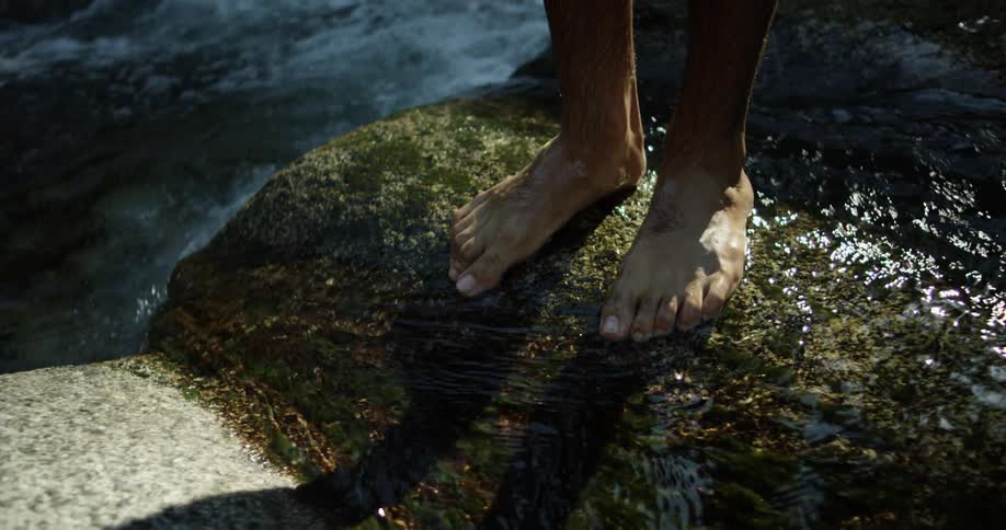 A man stands barefoot on a moss covered rock in a river