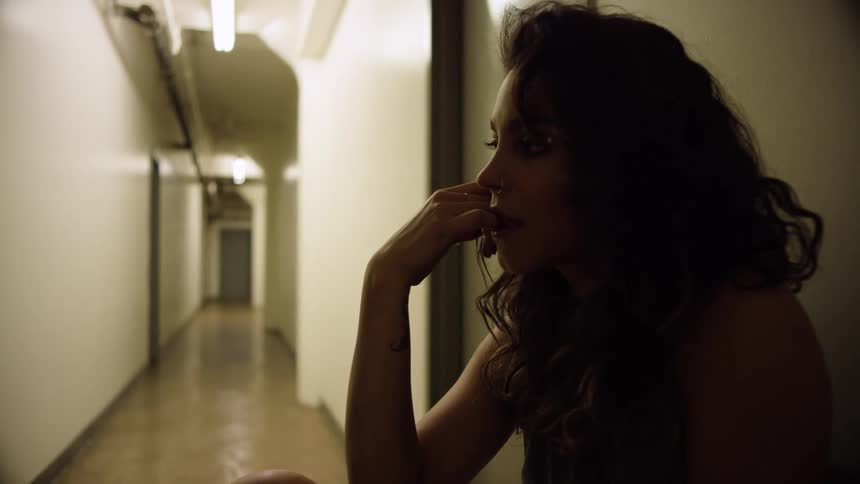 A woman sits in a hallway with her hand to her face and looking depressed