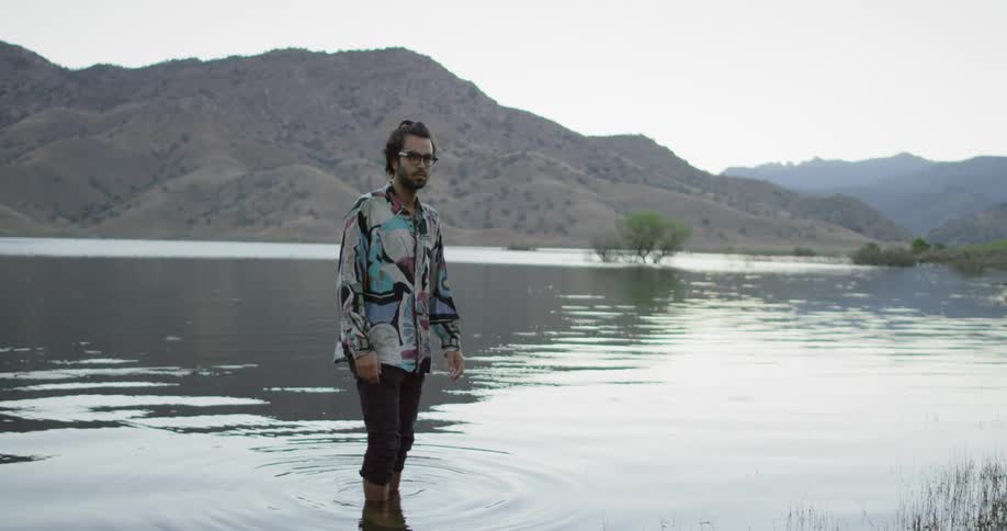 A hipster with his pants rolled up stands in a lake causing ripples as he turns