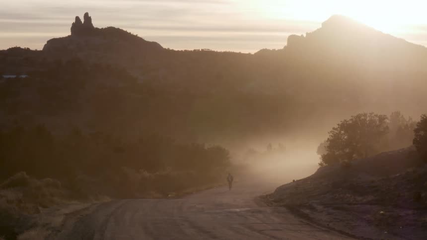 A person is walking down a dirt road away from a desert mountain in the evening
