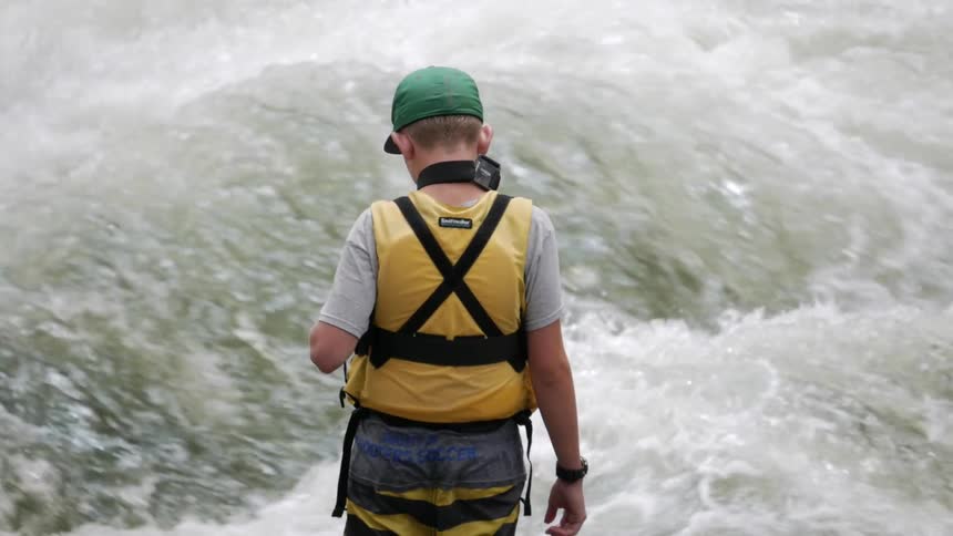 A boy in a yellow life vest stands near choppy, white water