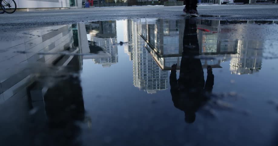A person walking on a wet street with a puddle that has reflections of buildings