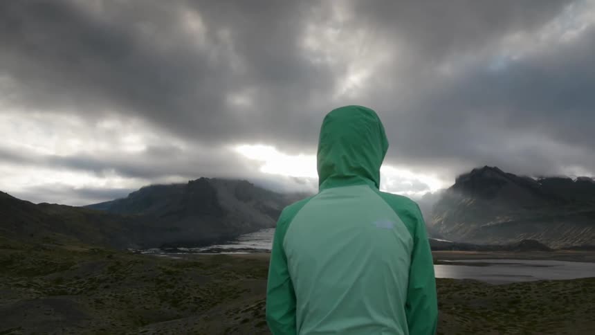 A person is standing before a small body of water by a mountain on a cloudy day