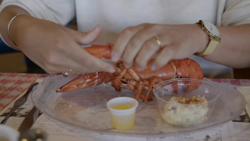 A woman sitting at a table in a restaurant pulls the claw off of a lobster and breaks it apart