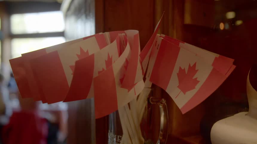 A bunch of small canadian flags in a clear glass mug with people sitting at a table by the window
