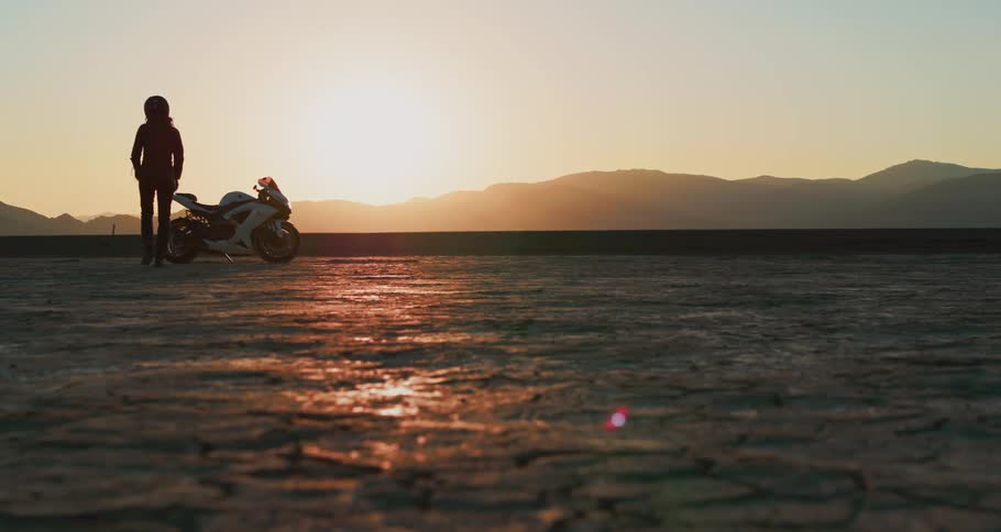 Female wearing a helmet walks toward a motorcycle sitting in the middle of the desert as the sun is setting while putting on a pair of gloves