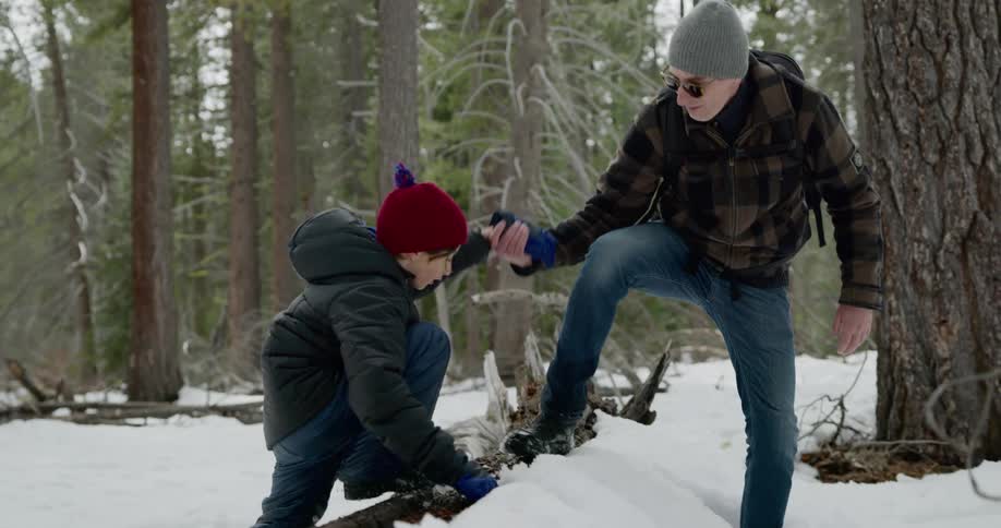 A man helps his son climb over a log while they are hiking in the snow covered forest