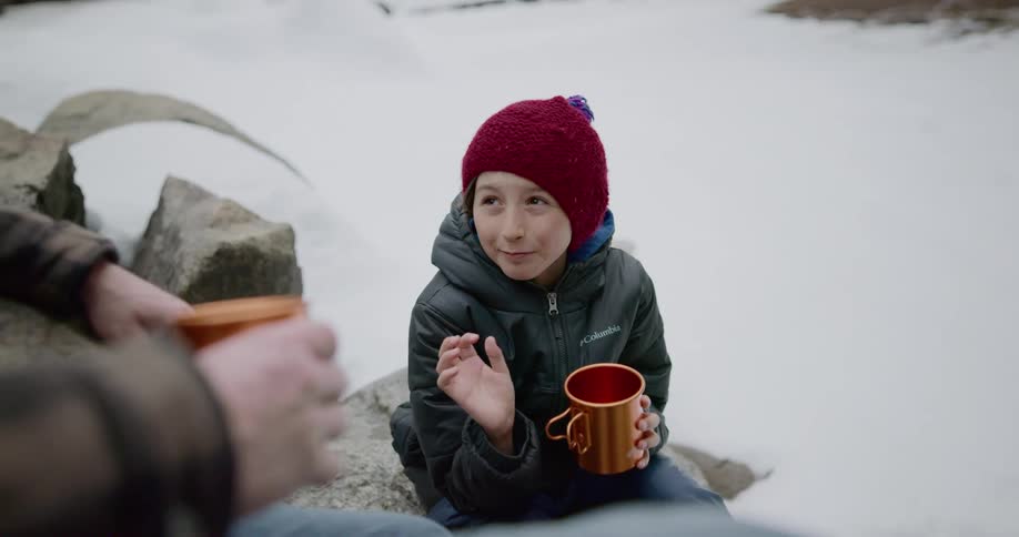 A child sits on a rock in a snow covered terrain holding a mug and talking to an adult